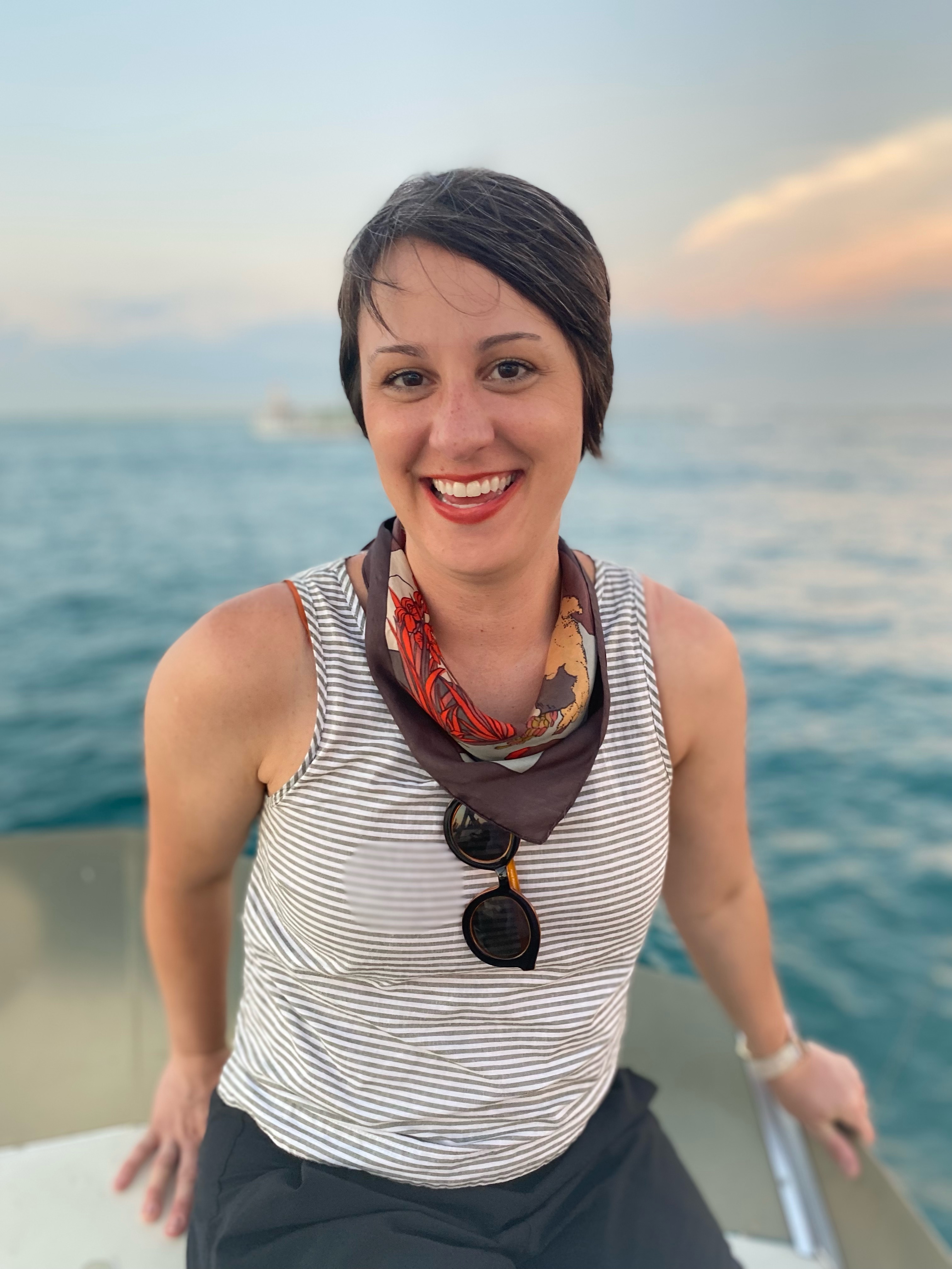 photo of Jacqueline Kish, a young white woman with short brown hair. She is wearing a striped tank top and a bandana around her neck with sunglasses and is wearing blue pants. She is sitting on a boat with water behind her.