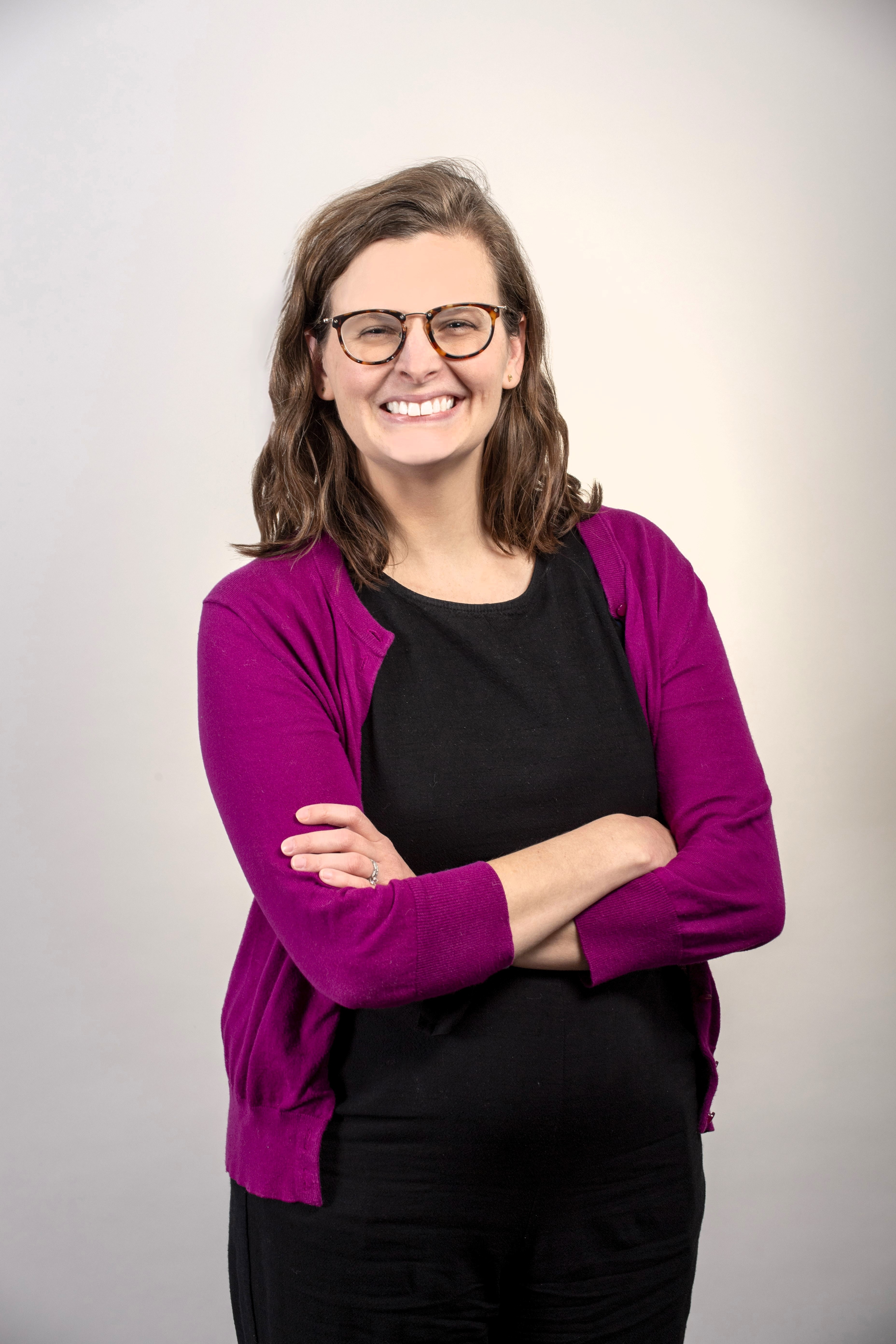 Photo of Amy McArthur, a young white woman with shoulder length brown hair wearing black pants and a black shirt with a pink cardigan. She has glasses and is crossing her arms and smiling.