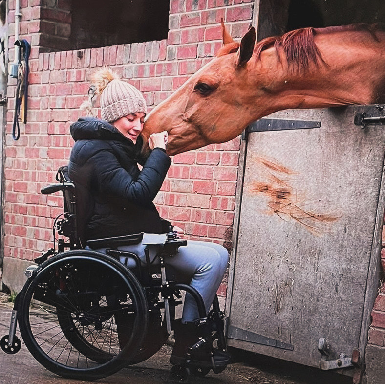 photo of Sarra, a young white woman in a wheelchair in a coat and hat nuzzling a brown horse