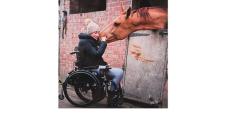 a photo of Sarra, a young woman with brown hair in a wheelchair giving a brown horse a nuzzle on the muzzle