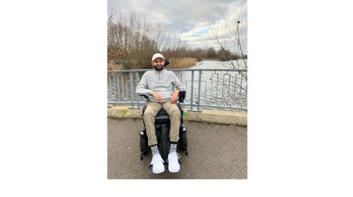 color photo of Viraj, a young man of Indian descent, sitting in a wheelchair outdoors on a bridge with a stream below.