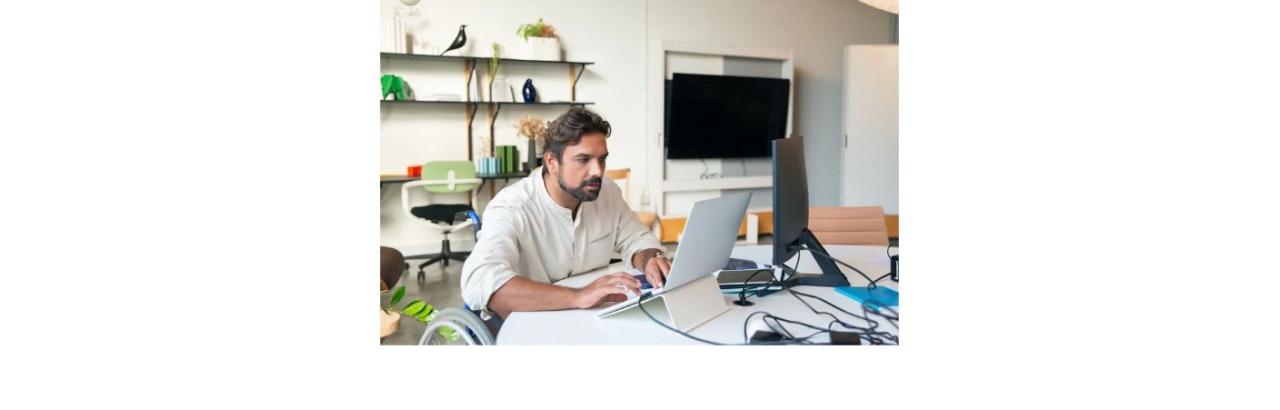 A white man with brown hair and beard sitting in a wheelchair works at a computer.