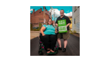 A person with a disability is sitting in a wheelchair while standing next to someone holding a green sign that reads "We are all human" in English and Spanish. 